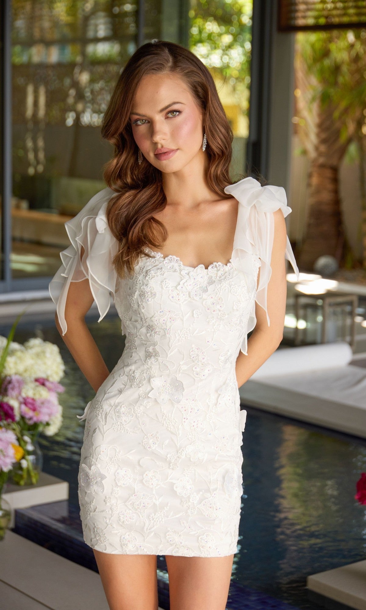 A woman stands indoors wearing the Ava Presley 47307 short homecoming dress featuring white floral embroidery and ruffled sheer sleeves; a pool and flowers are visible in the background.